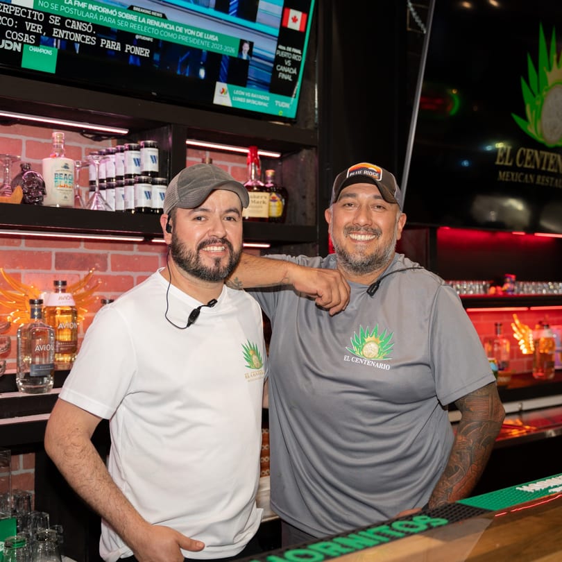 Two smiling men in El Centenario shirts stand behind a bar with liquor bottles and a TV screen.