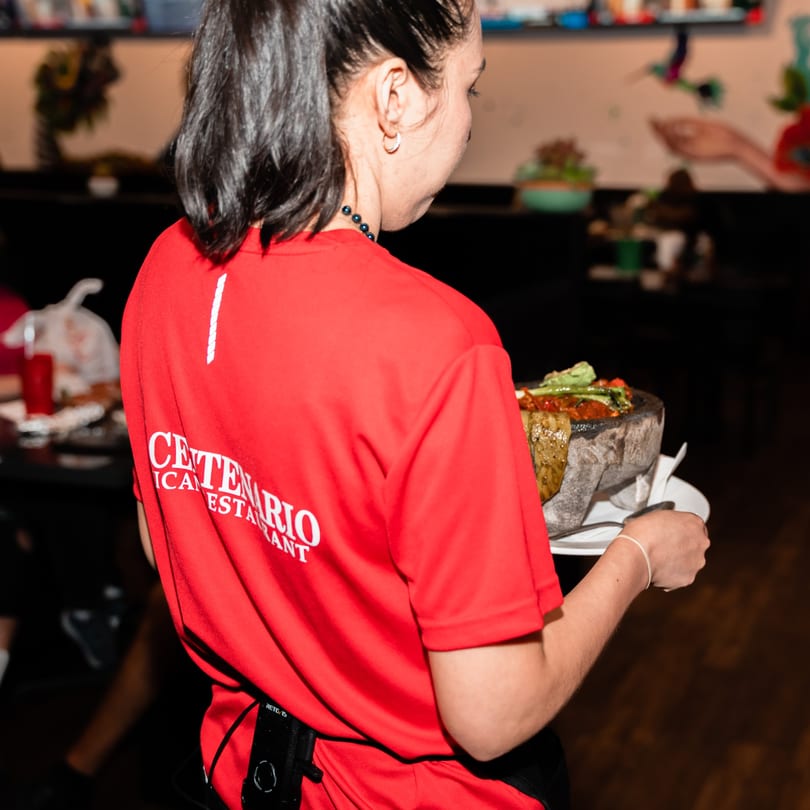 A server in a red El Centenario shirt carries a large molcajete filled with Mexican food.