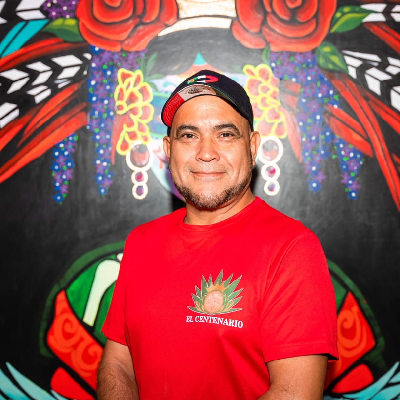 Man in a red El Centenario shirt and cap smiles against a colorful Mexican-themed mural.