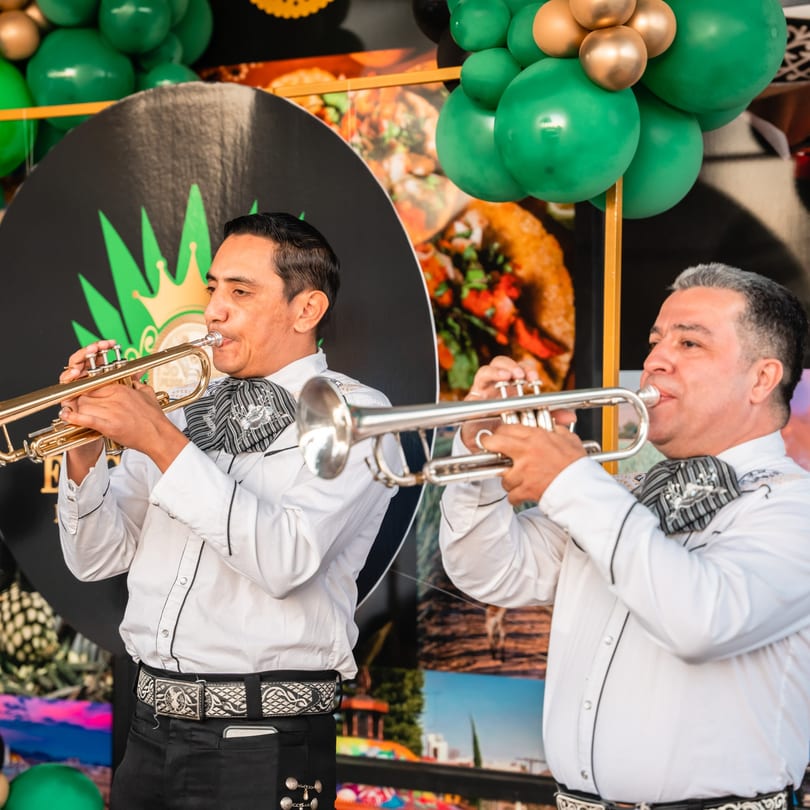 Two mariachi musicians in white shirts and charro ties playing trumpets at El Centenario WPB.