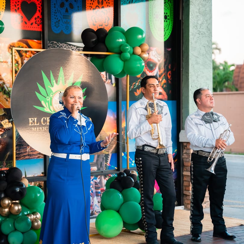 A woman in a blue mariachi dress sings into a microphone next to two mariachi trumpet players.