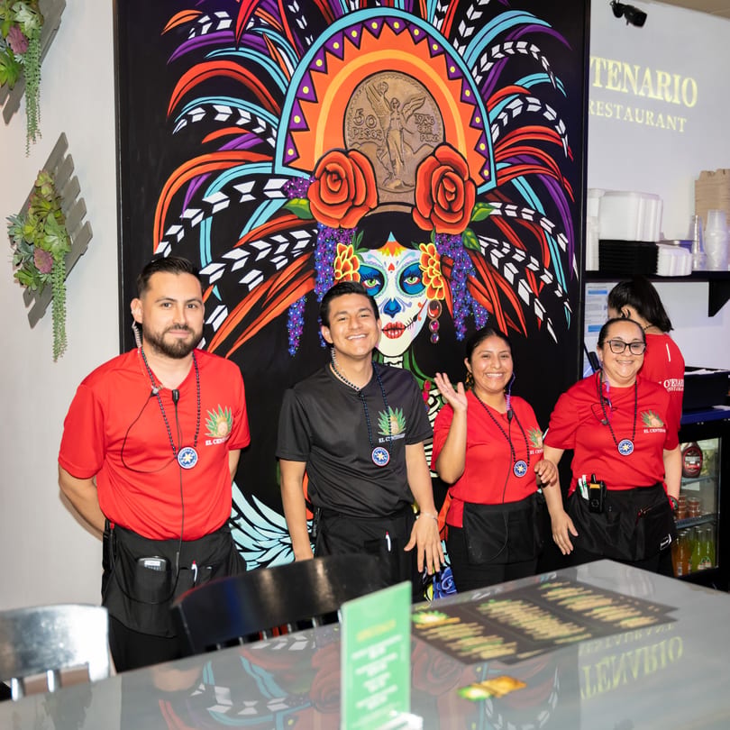 Four El Centenario WPB staff members in red and black uniforms stand in front of a colorful mural.