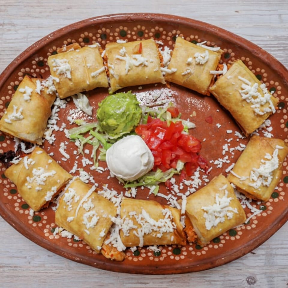 A platter of El Centenario WPB's Mexican flautas, topped with cheese, guacamole, sour cream, and diced tomatoes.