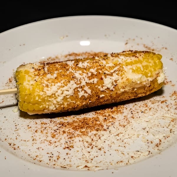 Elote, Mexican street corn, on a stick, covered in cheese and chili powder on a white plate.