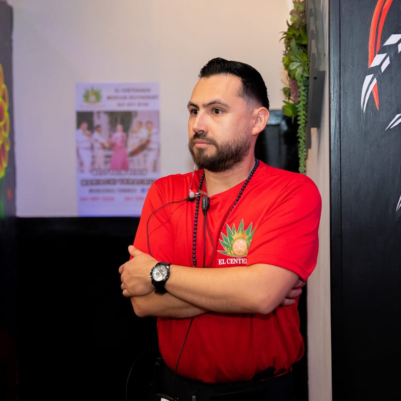 A man in a red El Centenario WPB shirt with a headset stands with crossed arms.