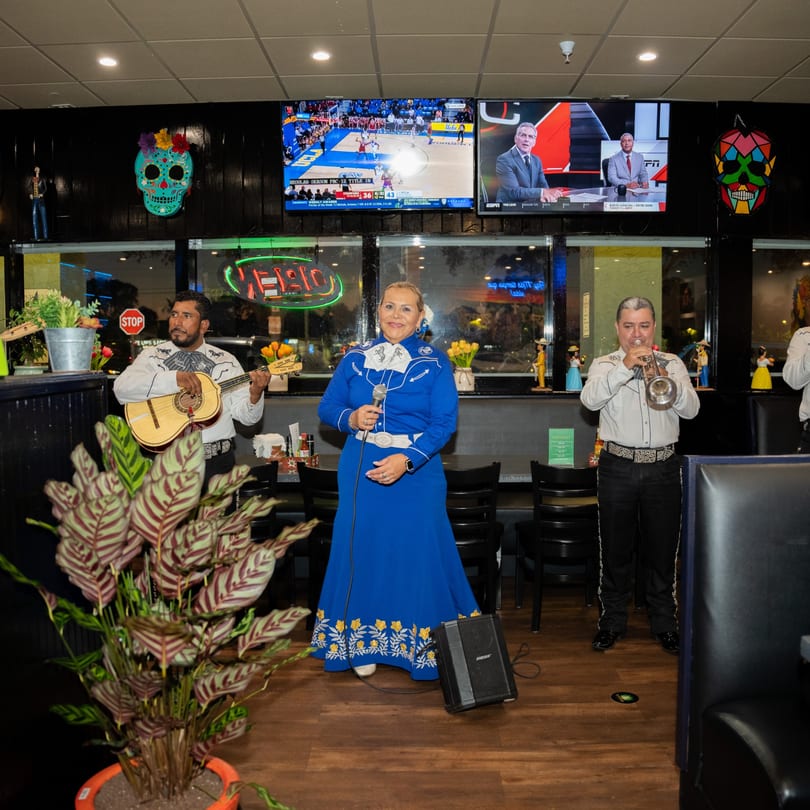 Mariachi band performing at El Centenario WPB, with a female singer in a blue dress and two male musicians.
