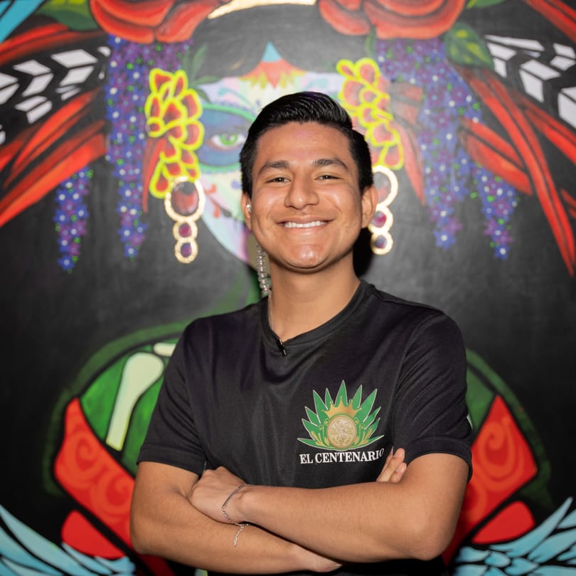Smiling man in an El Centenario shirt with arms crossed, in front of a colorful mural.