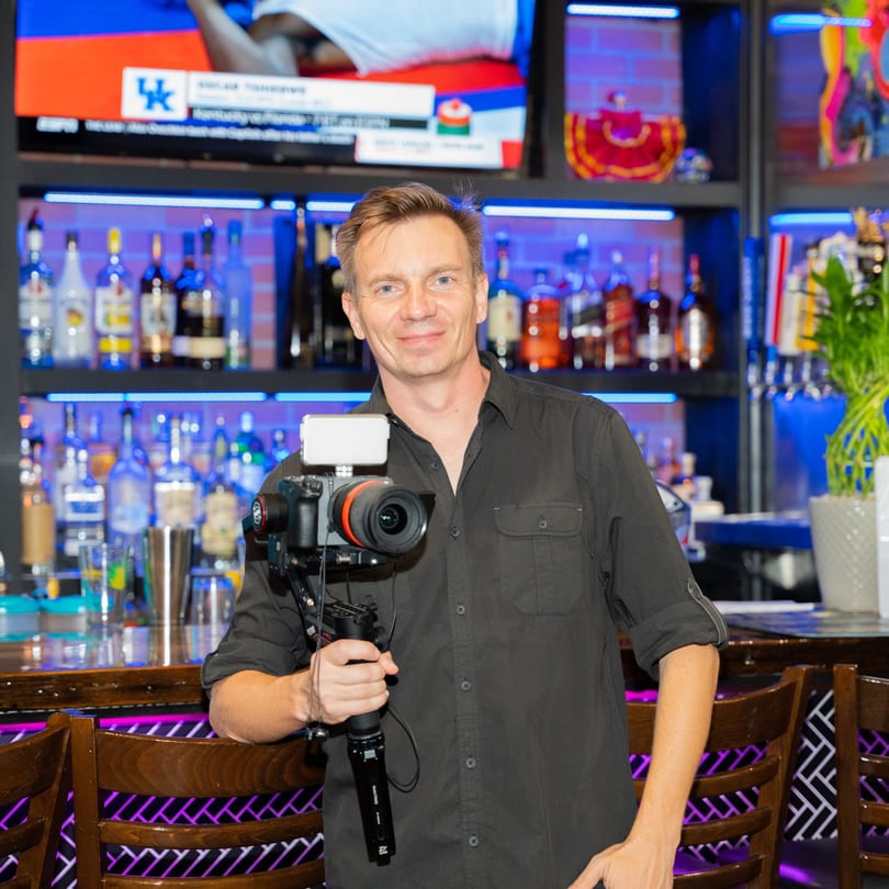 A man holding a camera on a gimbal stands in front of a bar with bottles and neon lights.