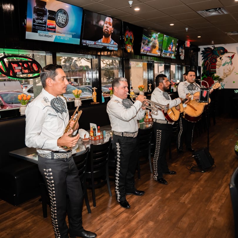 Four mariachi musicians in traditional attire perform with instruments inside El Centenario WPB restaurant.
